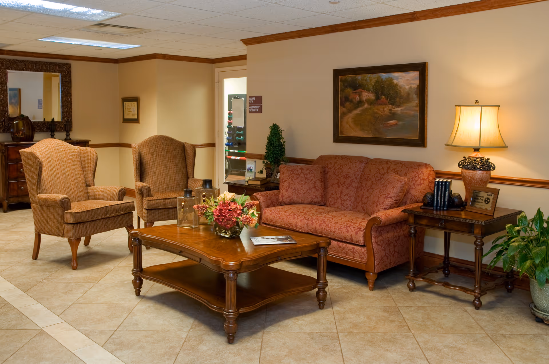 A cozy living room area with a patterned red sofa, two brown upholstered armchairs, a wooden coffee table with a floral centerpiece, and a side table with a lamp and decorative items. The room has beige tiled flooring, cream-colored walls with wooden trim, a framed landscape painting, and a potted plant in the corner.