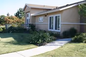 Single-story beige stucco apartment building with a paved walkway, lawn, and landscaped shrubs.