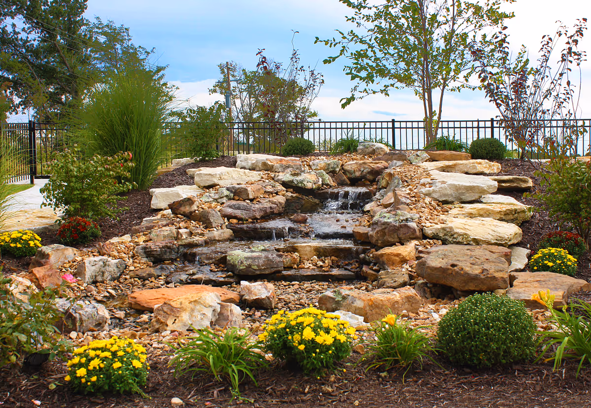 A landscaped outdoor garden area featuring a small cascading rock waterfall surrounded by various green shrubs, yellow flowers, and other plants, with a black metal fence and trees in the background under a partly cloudy sky.