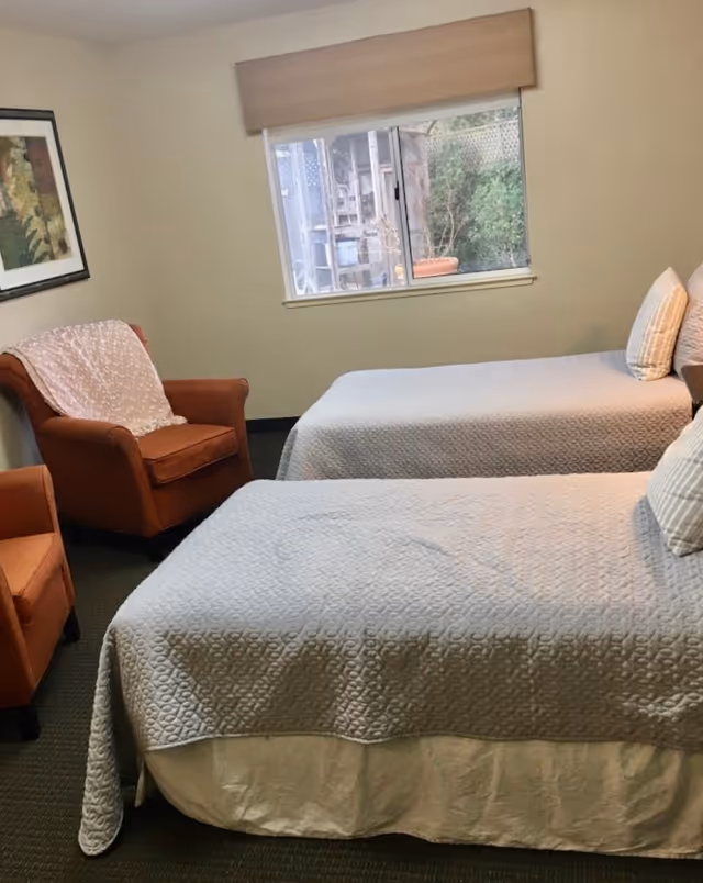 Small bedroom with two twin beds covered in light gray quilts, two orange armchairs, a framed picture, and a window.