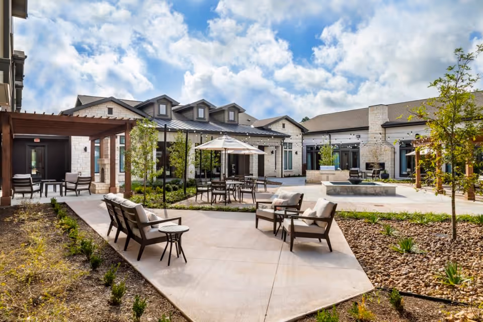 Outdoor courtyard with patio seating, umbrellas, pergolas, landscaped beds, and a central fountain in front of a low-rise building under a partly cloudy sky.