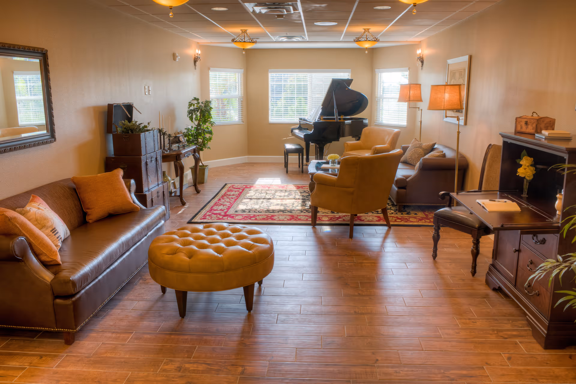 A cozy living room with wooden flooring, a brown leather sofa with orange cushions, a round tufted ottoman, two armchairs, a glass coffee table, a grand piano near three windows, a patterned area rug, a wooden desk with a chair, and warm lighting from ceiling fixtures and floor lamps.