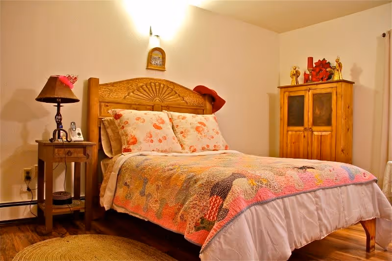 A cozy bedroom with a wooden bed frame featuring a carved headboard. The bed is made with floral pillows and a colorful patchwork quilt. To the left of the bed is a wooden nightstand with a lamp, a phone, and a small decorative item. On the right side of the room, there is a rustic wooden cabinet with decorative items on top. The room has light-colored walls and wooden flooring with a round woven rug partially visible.
