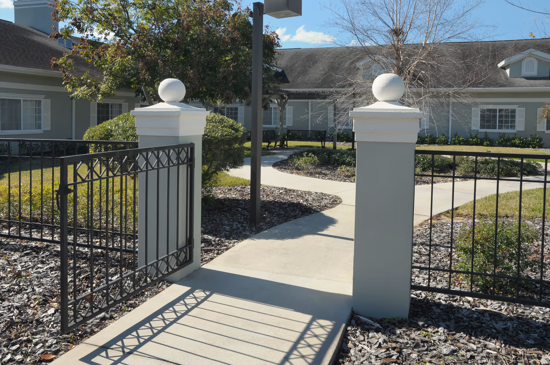 Open black metal gate with white pillars leading to a curved concrete walkway surrounded by landscaped bushes and trees, with a single-story building in the background under a clear blue sky.