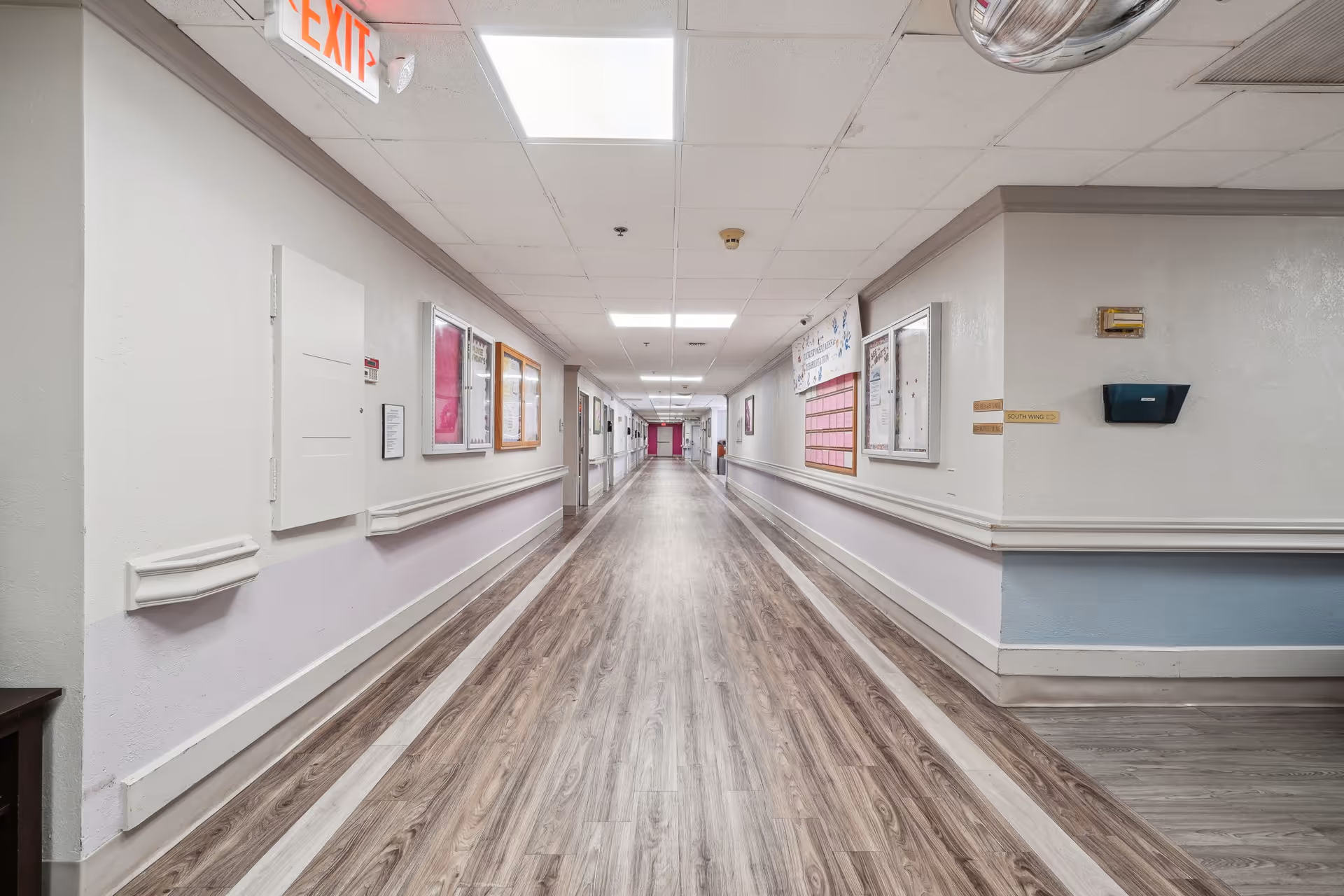 Long, well-lit hallway in a nursing and rehab center with wood-patterned flooring, white walls with a light purple lower section, handrails on both sides, bulletin boards and signs on the walls, and an exit sign on the ceiling.