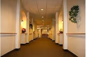 A long, well-lit hallway in an assisted living facility with beige walls, brown carpet, and decorative plants and flowers on ledges along the walls.