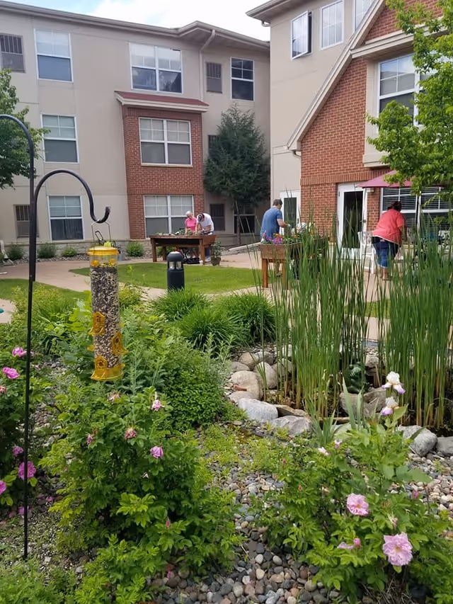 Outdoor garden area at The Pines Senior and Assisted Living with green plants, pink flowers, a bird feeder, and a small pond surrounded by rocks. In the background, three people are tending to plants on raised garden beds near a beige and red brick building.