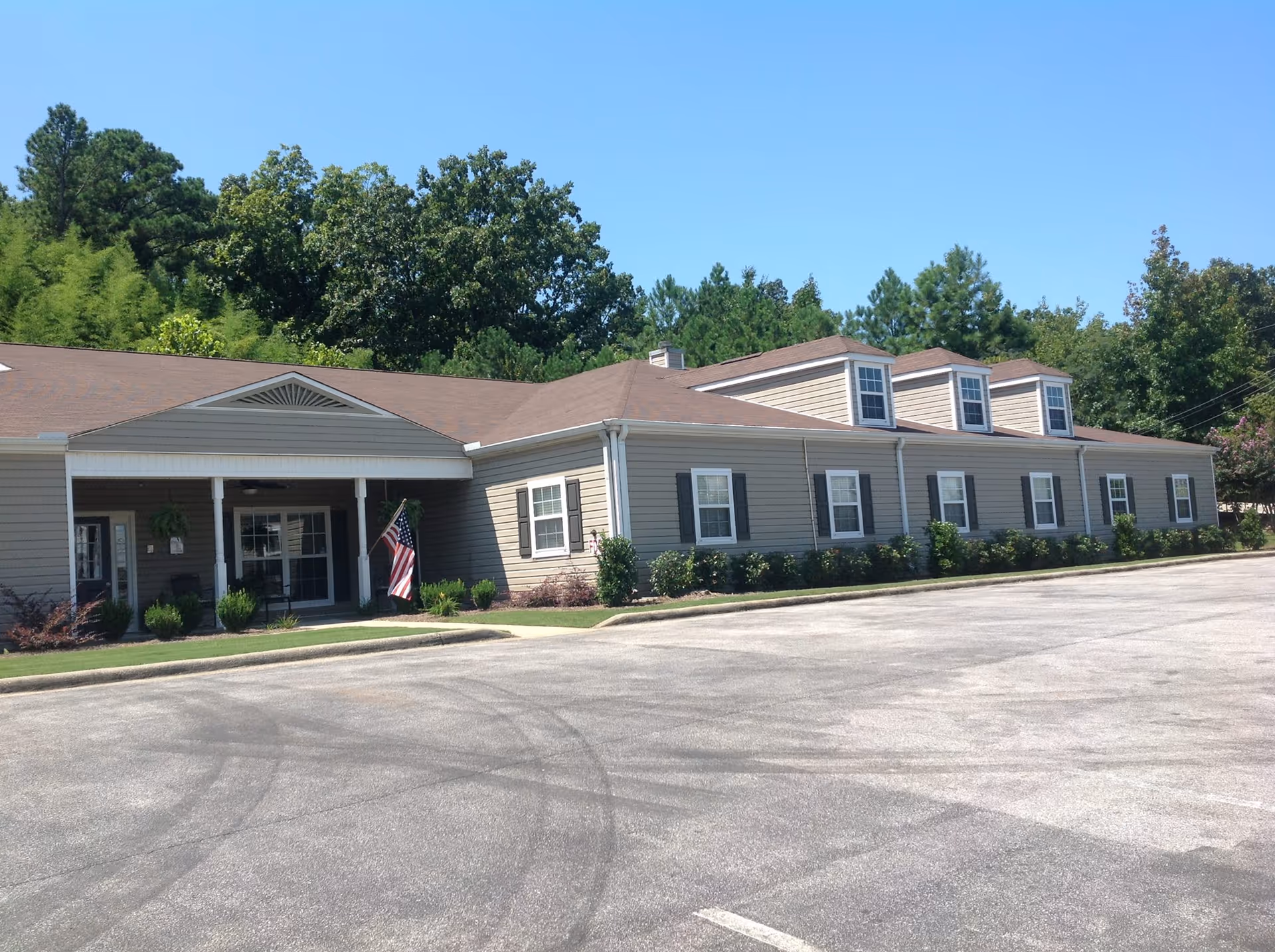 Exterior view of a single-story building with beige siding and a brown roof, surrounded by greenery and trees under a clear blue sky. There is a small covered entrance with white columns and an American flag displayed near the door. The foreground shows an empty parking lot.