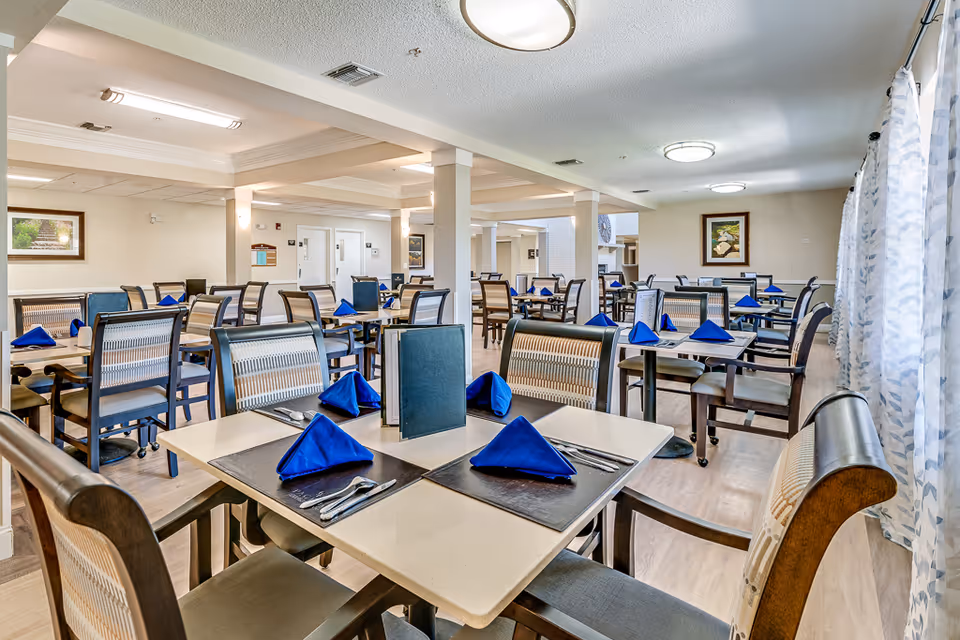A spacious dining room in a senior living facility with multiple tables set with blue folded napkins, menus, and silverware. The room has light-colored walls, large windows with patterned curtains, framed artwork on the walls, and ceiling lights providing bright illumination.