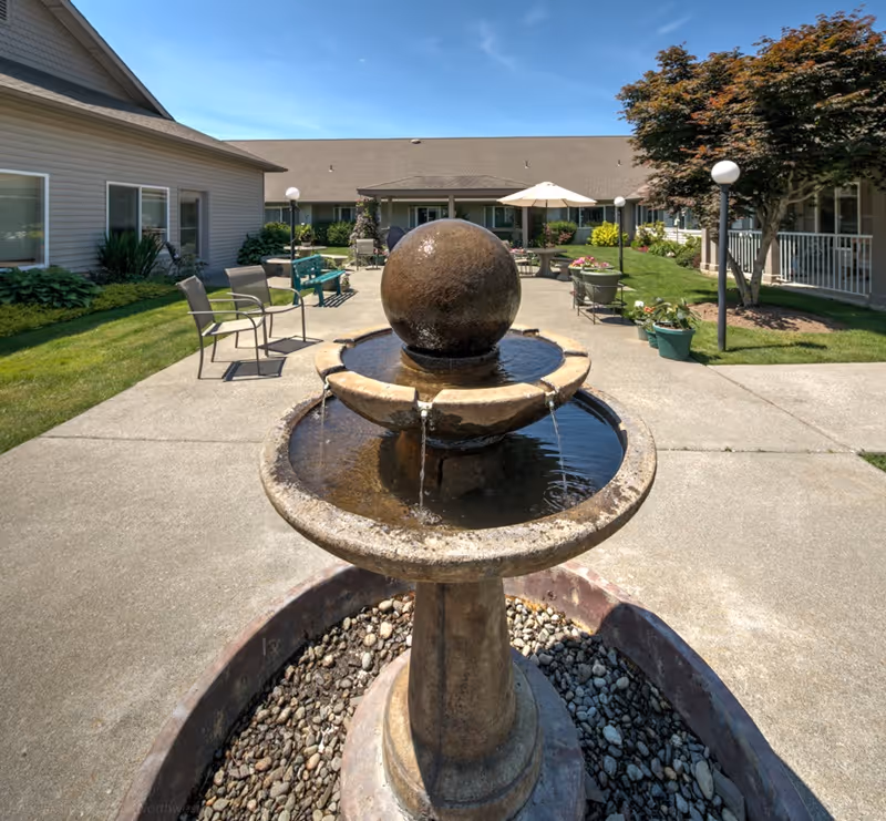 Outdoor courtyard area at a senior living facility featuring a stone water fountain with a spherical top, surrounded by a circular basin with flowing water. The courtyard has concrete walkways, green grass, potted plants, outdoor chairs, benches, and a patio umbrella. The buildings surrounding the courtyard have beige siding and windows, with a clear blue sky overhead.