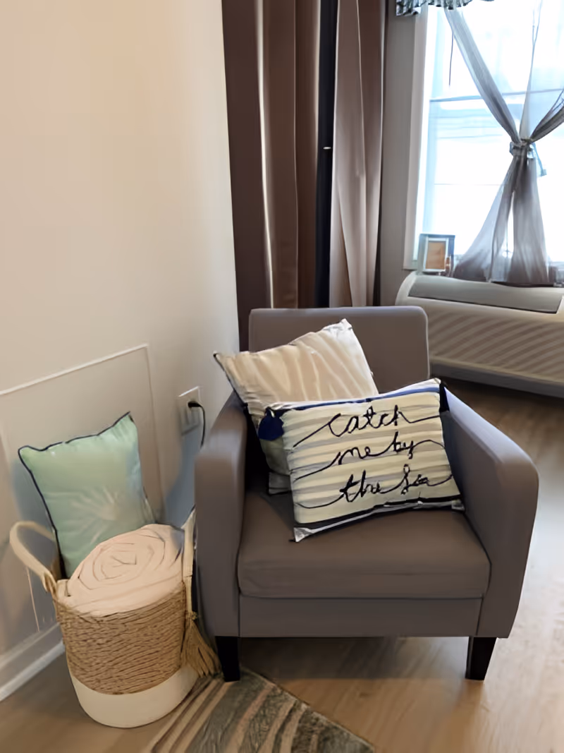 A cozy corner with a gray armchair adorned with two decorative pillows, one with the text 'catch me by the sea'. Next to the chair is a woven basket containing a rolled blanket and a light green pillow. In the background, there is a window with sheer curtains and a heating or air conditioning unit beneath it.