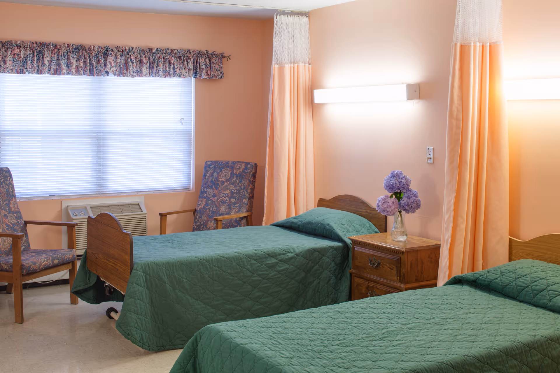 A room with two single beds covered in green quilts, separated by a wooden nightstand with a vase of purple flowers. The walls are peach-colored with peach and white privacy curtains hanging between the beds. Two floral-patterned armchairs are placed near a window with blinds and a floral valance. A wall-mounted light fixture is above each bed.