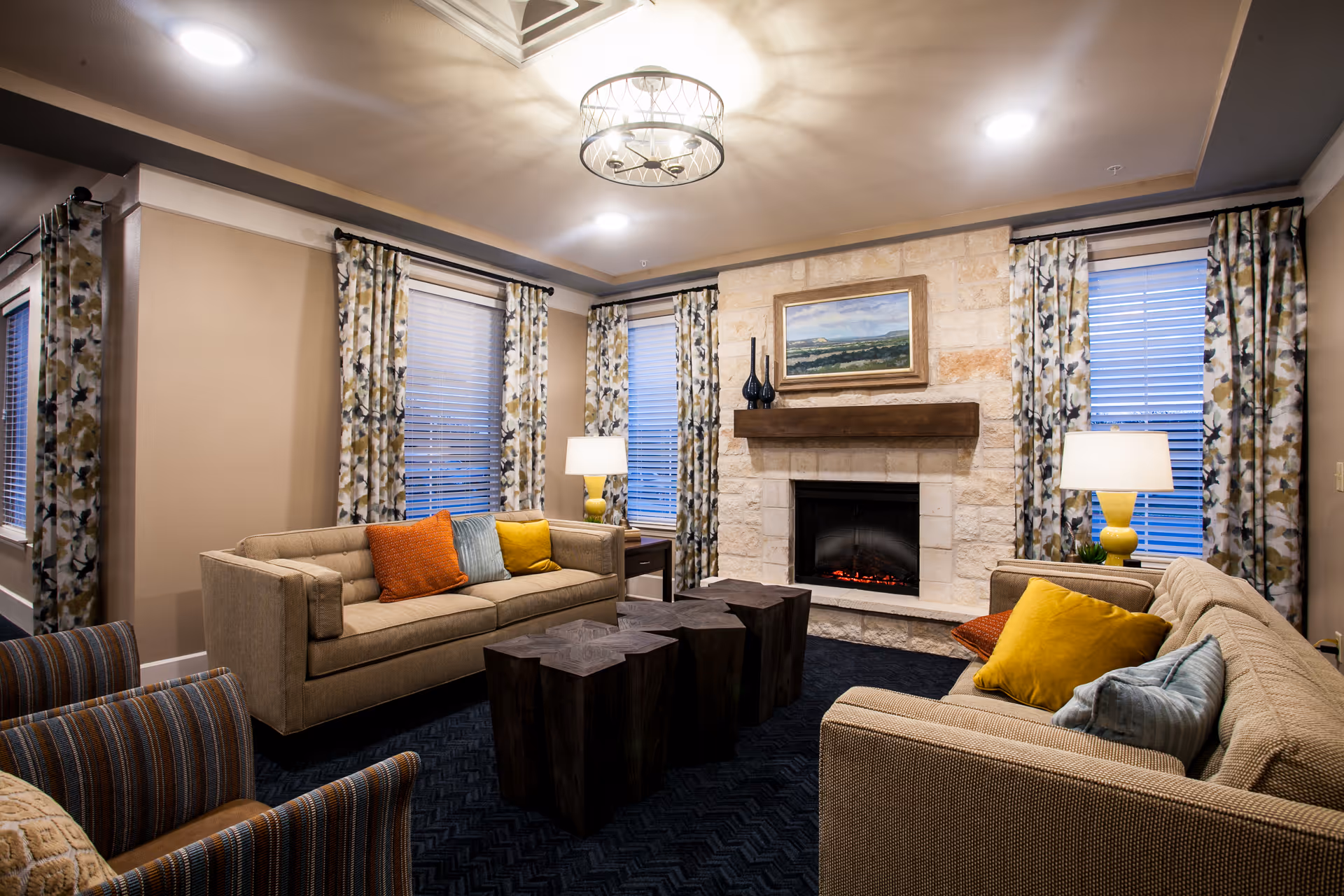 A cozy living room with two beige sofas adorned with colorful throw pillows in orange, yellow, and blue. There are three dark wooden coffee tables in the center. The room features a stone fireplace with a wooden mantel, above which hangs a landscape painting. The walls are beige with floral patterned curtains on the windows, and two table lamps with yellow bases provide warm lighting.