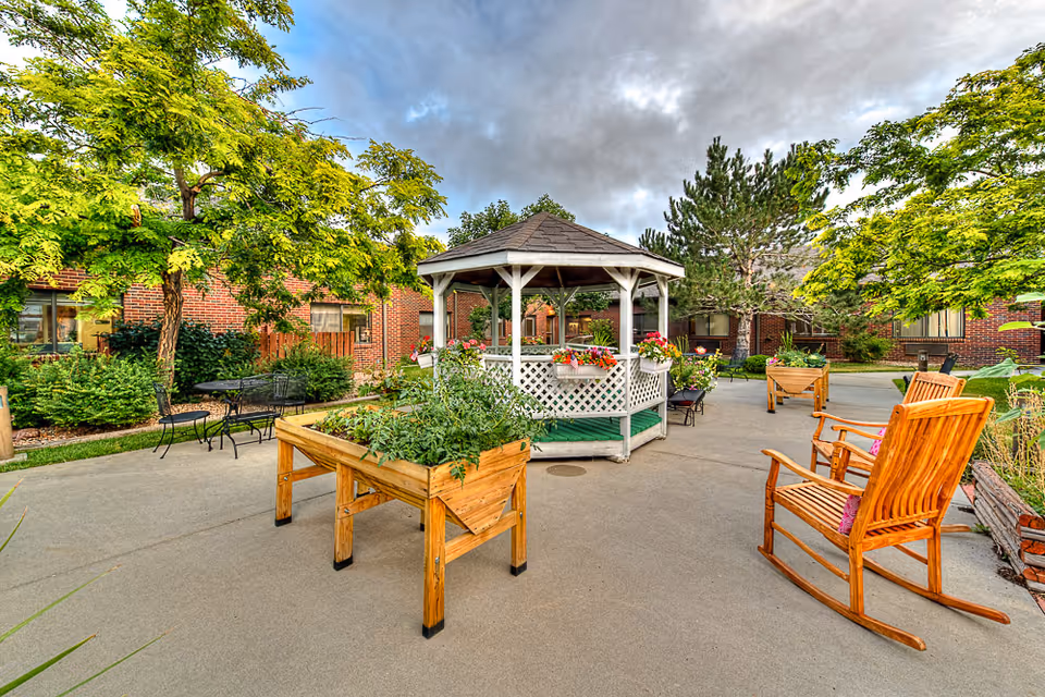 Outdoor courtyard with a white gazebo, raised wooden planters, rocking chairs, and surrounding brick building.