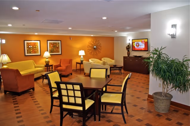 A cozy living room area with a wooden table and four chairs with yellow cushions in the foreground. The background features a seating area with two orange armchairs, a yellow sofa, two beige armchairs, side tables with lamps, wall art, a decorative wall piece, a flat-screen TV mounted on a cabinet, and a potted plant near the wall.