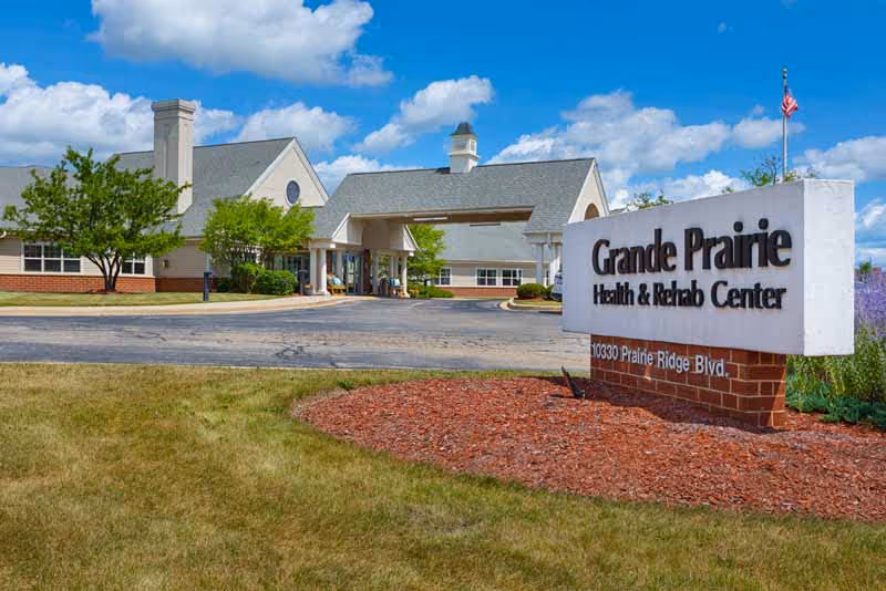 Exterior view of Grande Prairie Health and Rehabilitation Center building with a large sign displaying the facility name and address. The building has a covered entrance, surrounded by trees and landscaping under a partly cloudy blue sky.
