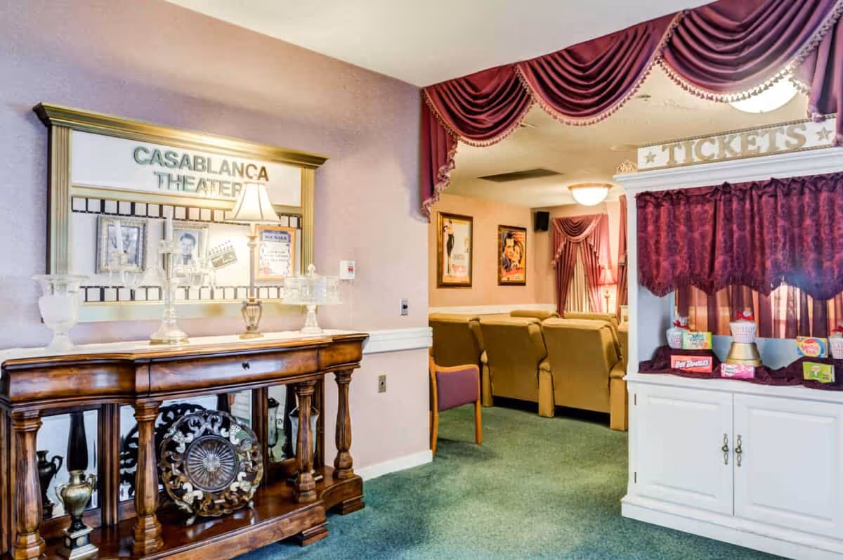 Interior view of Casablanca Theater at Newhaven Court at Clearview featuring a ticket booth with a burgundy curtain, a wooden console table with decorative items, and a seating area with beige recliners and movie posters on the walls.