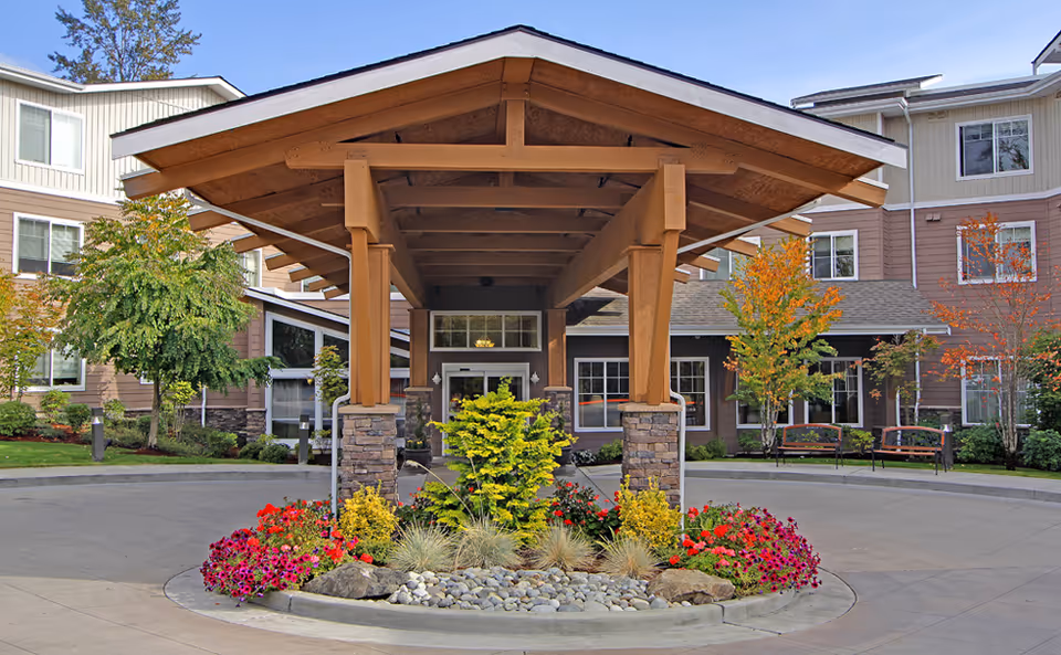 Entrance of Radcliffe Place senior living facility with a covered driveway supported by wooden beams and stone pillars. The building is surrounded by landscaped gardens with colorful flowers, shrubs, and trees showing autumn colors. Benches are visible near the building's windows.