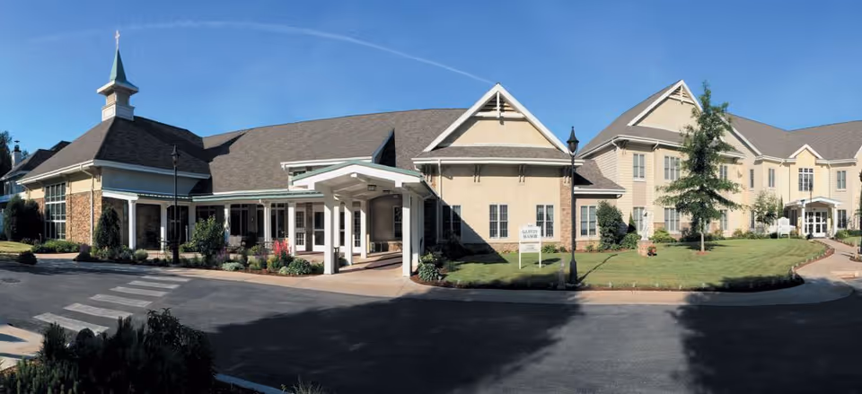 Exterior view of Garvey Manor Nursing Home building with a driveway, landscaped lawn, and a clear blue sky. The building features a covered entrance, multiple windows, and a small steeple with a cross on top.