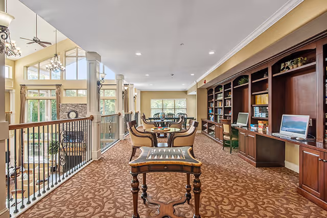 Interior view of a senior living facility common area with patterned carpet, wooden bookshelves filled with books and computers on the right, several tables and chairs arranged for seating, and a railing overlooking a lower level with large windows and a stone fireplace.