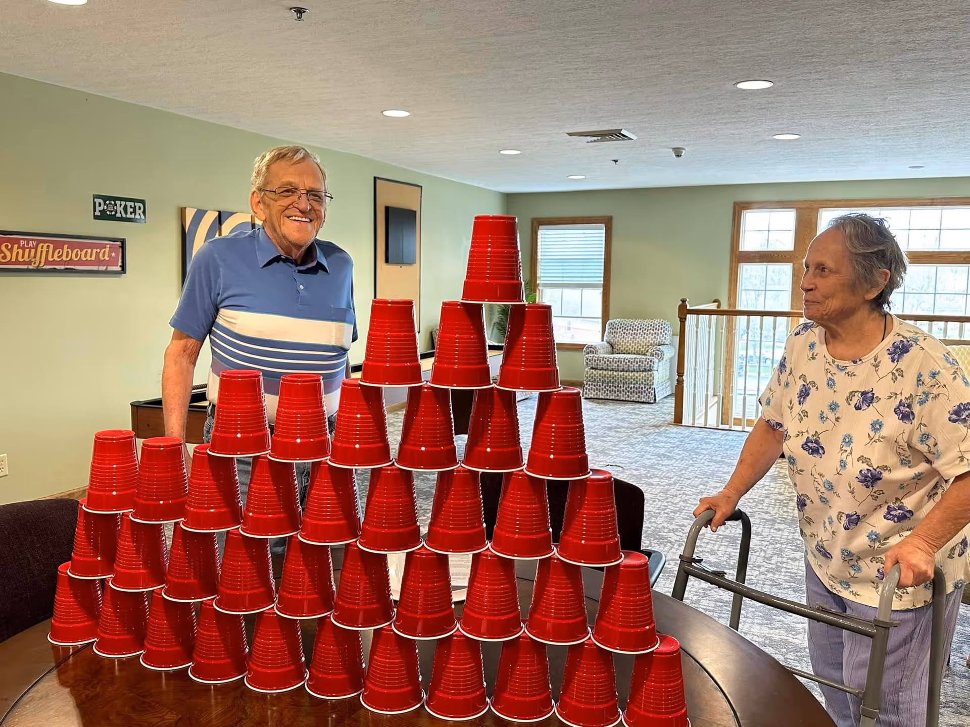 An elderly man and woman in a common area of a senior living facility stand near a table with a large pyramid made of stacked red plastic cups. The man is smiling and wearing a blue striped polo shirt, while the woman, using a walker, wears a floral shirt. The room has light green walls, windows letting in natural light, and comfortable seating. Signs on the wall read 'Poker' and 'Play Shuffleboard'.