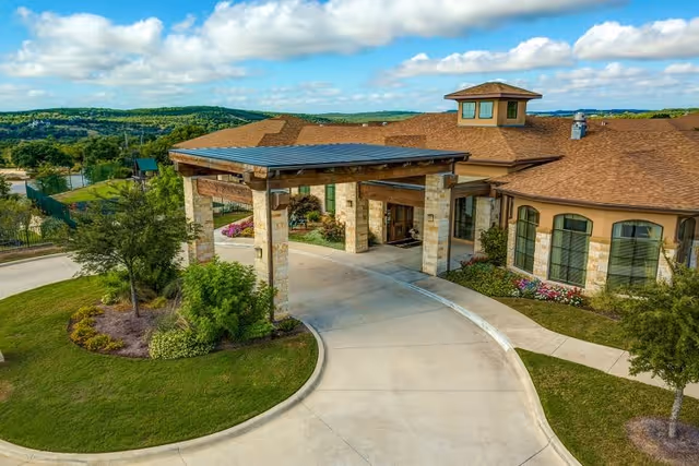 Exterior view of a senior living facility with a covered entrance supported by stone pillars, surrounded by landscaped greenery and trees under a partly cloudy sky.