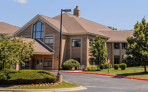 Exterior view of a two-story brick building with large windows, a peaked roof, and a covered entrance. The surrounding area includes a paved driveway, manicured bushes, trees, and a well-maintained lawn under a clear blue sky.