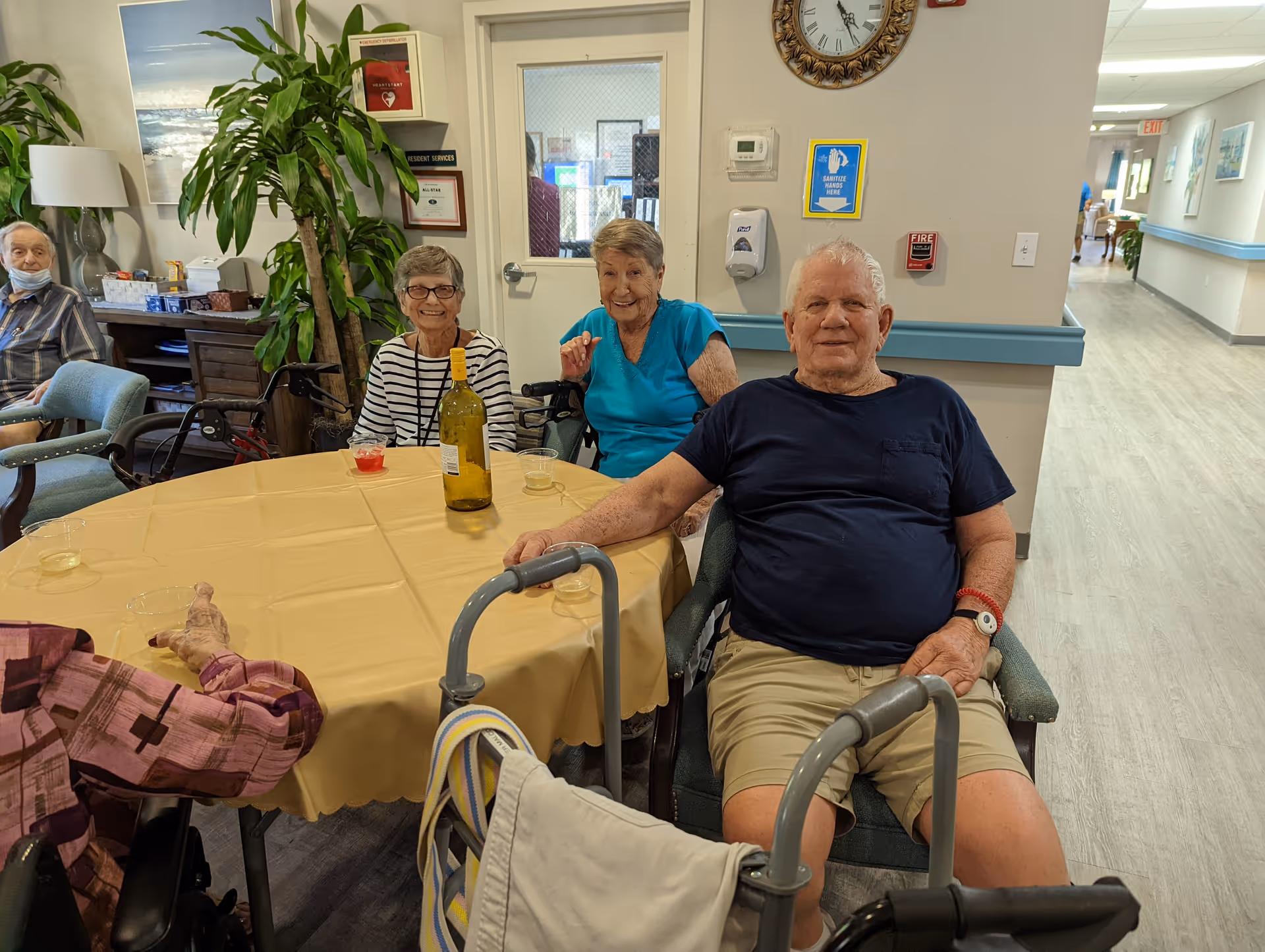 A group of elderly people sitting around a table with a yellow tablecloth in a common area of a senior living facility. There are drinks on the table, and a large plant and various wall decorations are visible in the background. The setting appears warm and social.
