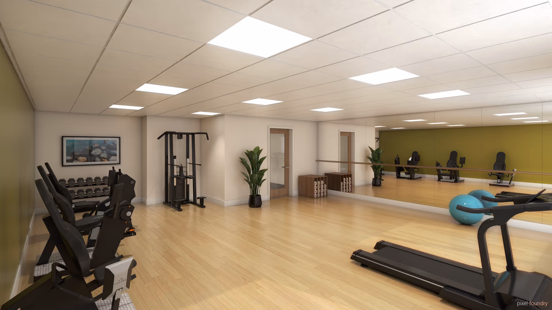 A spacious senior living facility exercise room with wooden flooring, several exercise machines including treadmills and weight machines, a large wall mirror, a ballet barre, two blue exercise balls, a plant, and a rack with rolled-up yoga mats. The room has a drop ceiling with fluorescent lighting and a framed picture on one wall.