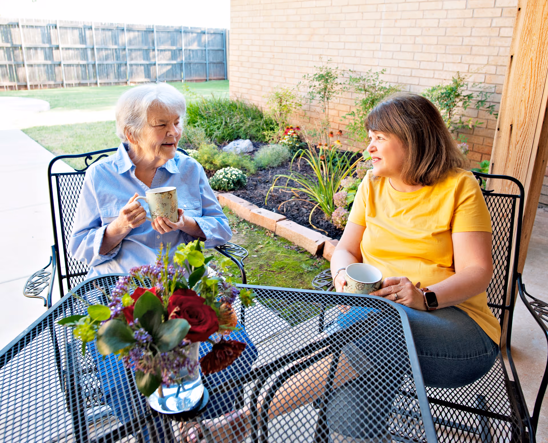 An elderly woman and a middle-aged woman sitting outdoors at a metal table with a flower vase, holding mugs and engaging in conversation. They are seated on metal chairs near a garden bed with plants and flowers, next to a brick wall and a wooden post.