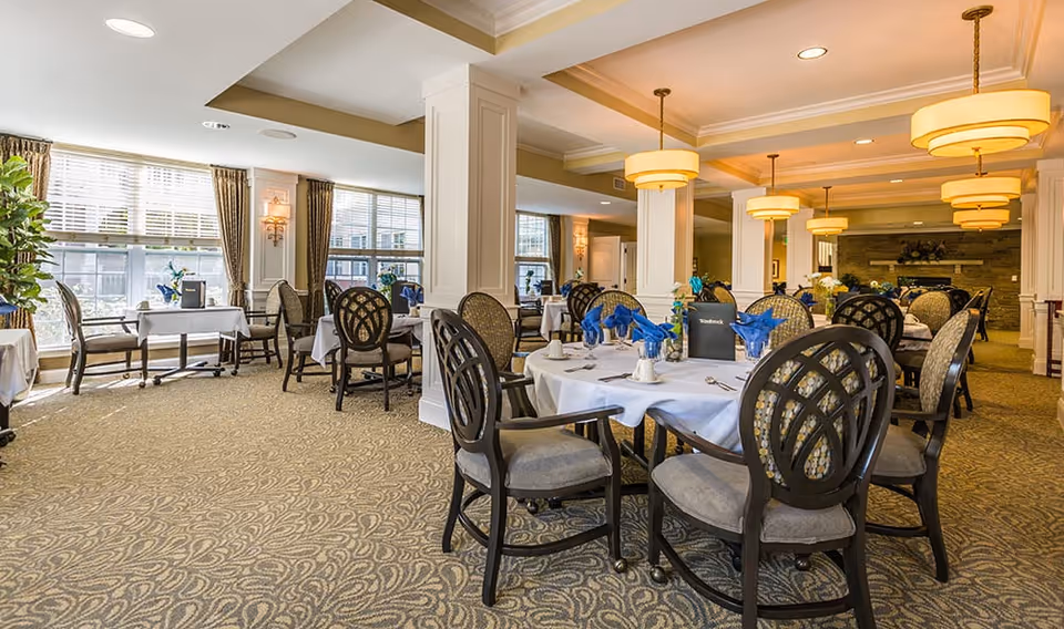 A spacious and well-lit dining room in a senior living facility with round tables covered in white tablecloths, each set with blue napkins, silverware, and small decorative items. The room features large windows with curtains, patterned carpet, elegant chairs, and warm ceiling lights.