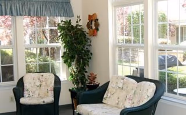 Sunlit sitting area with two cushioned wicker chairs, a small table and a potted plant in front of large windows.