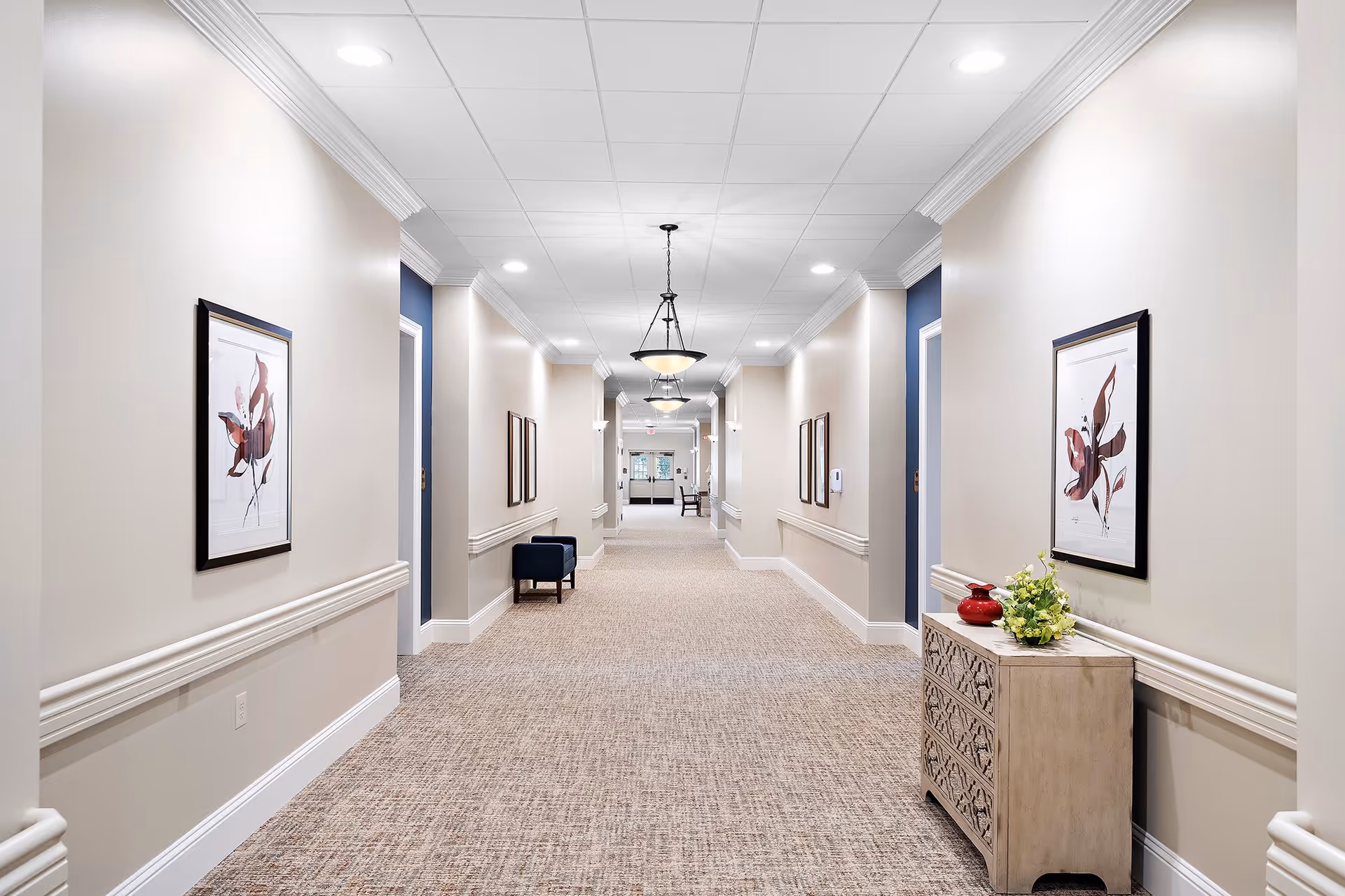 A well-lit, carpeted hallway in a senior living facility with beige walls, white crown molding, and blue door frames. The hallway features framed artwork on the walls, a small upholstered bench, and a decorative chest with a red vase and a flower arrangement. Ceiling lights and hanging lamps illuminate the corridor, leading to a door at the far end.