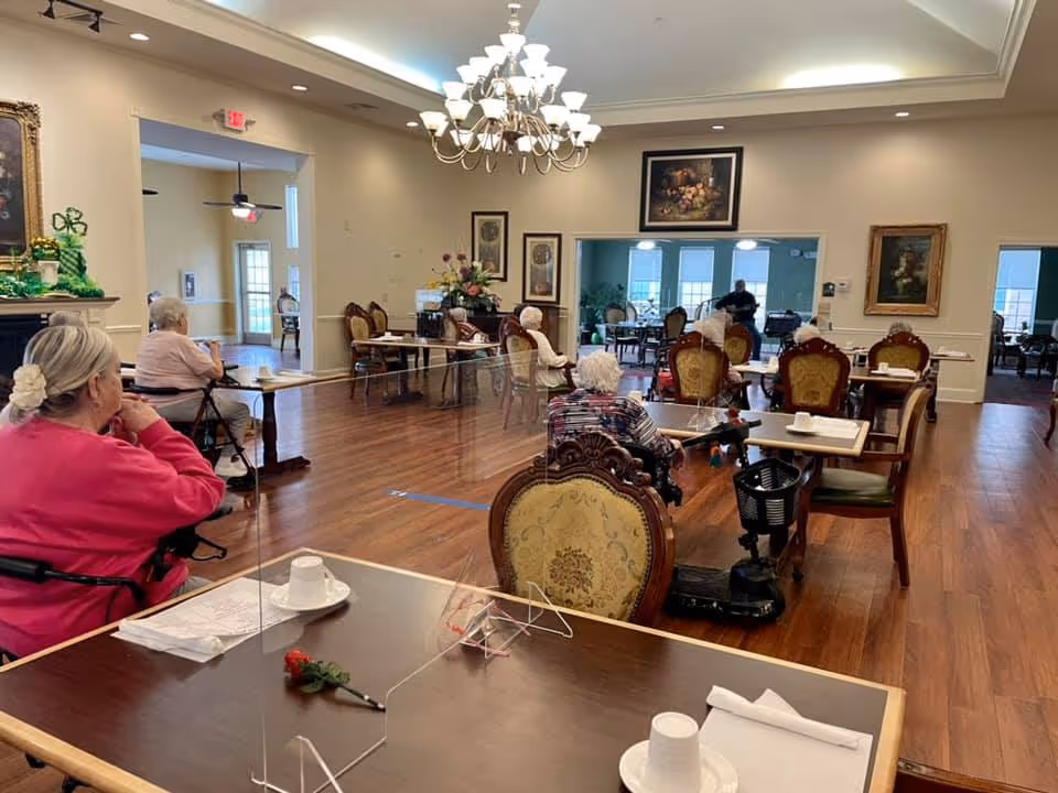 A dining room in a senior living facility with elderly residents seated at tables. The room has wooden floors, ornate chairs, and a chandelier hanging from the ceiling. Tables have white cups, napkins, and some have clear plastic dividers. Paintings and floral arrangements decorate the walls and room.
