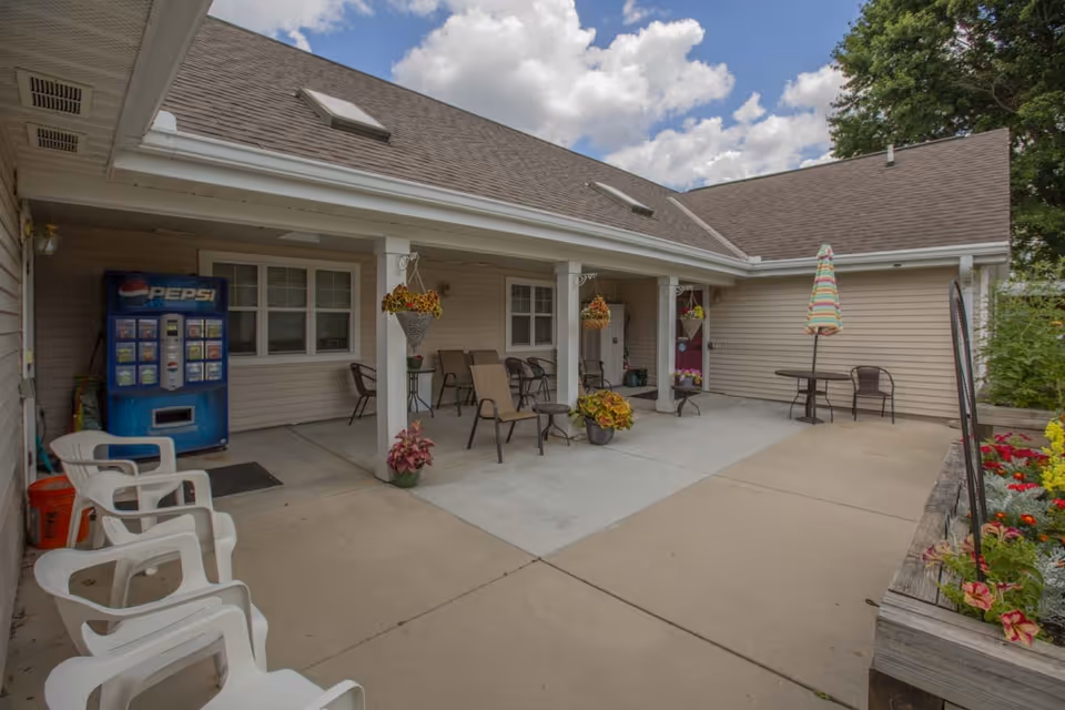 Outdoor patio area at Maple Meadows Assisted Living with several chairs, a Pepsi vending machine, hanging flower baskets, potted plants, a table with a colorful striped umbrella, and a garden bed with flowers on the right side under a partly cloudy sky.