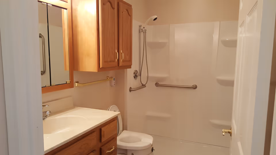 A bathroom with a white shower area equipped with grab bars and a handheld showerhead, a white toilet with a raised seat, a wooden vanity with a white countertop and sink, a wooden cabinet above the toilet, and a mirror above the sink.