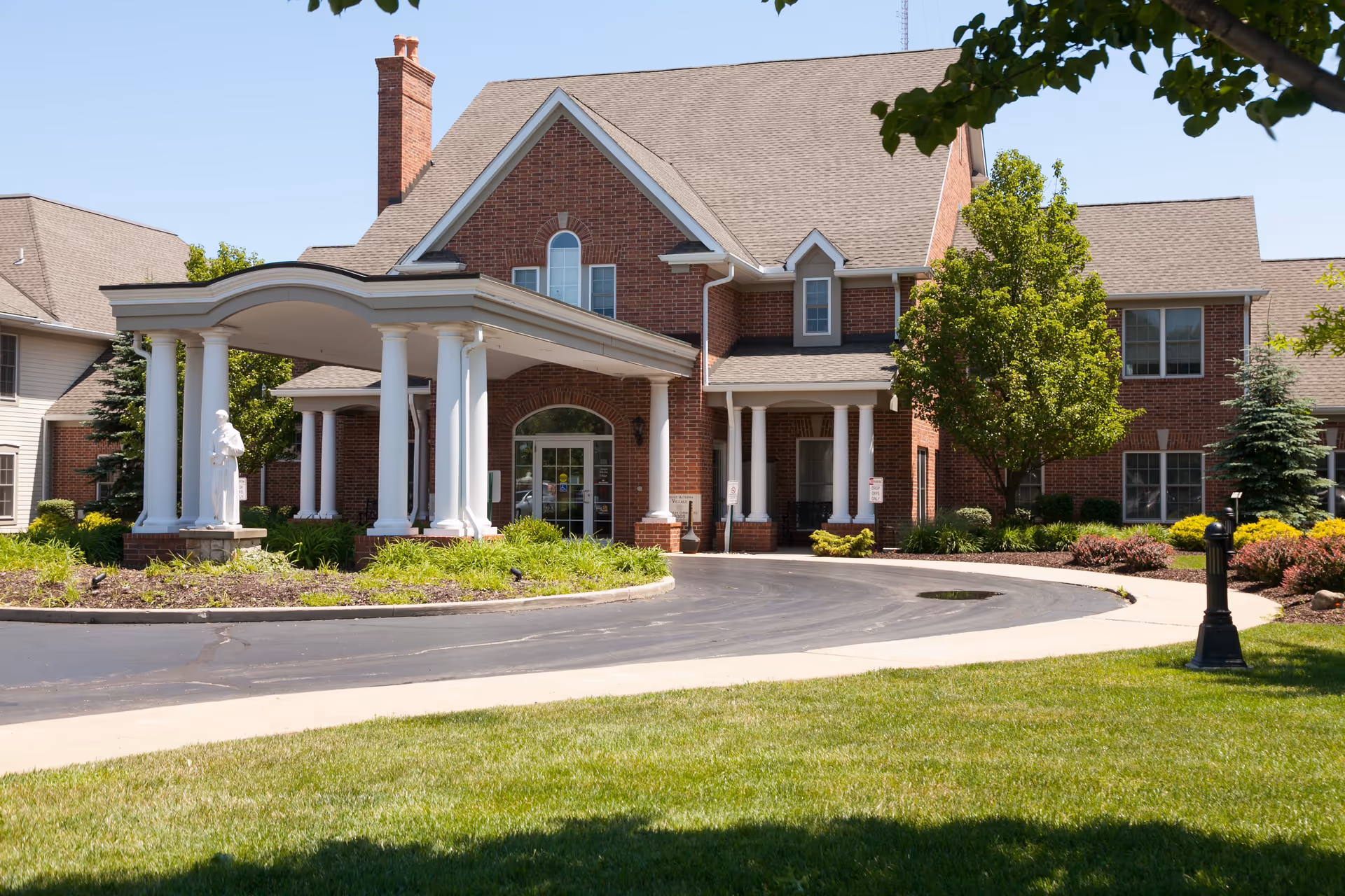 Front exterior view of Mount Alverna Village, a large brick building with a covered entrance supported by white columns, surrounded by green lawns, trees, and shrubs under a clear blue sky.