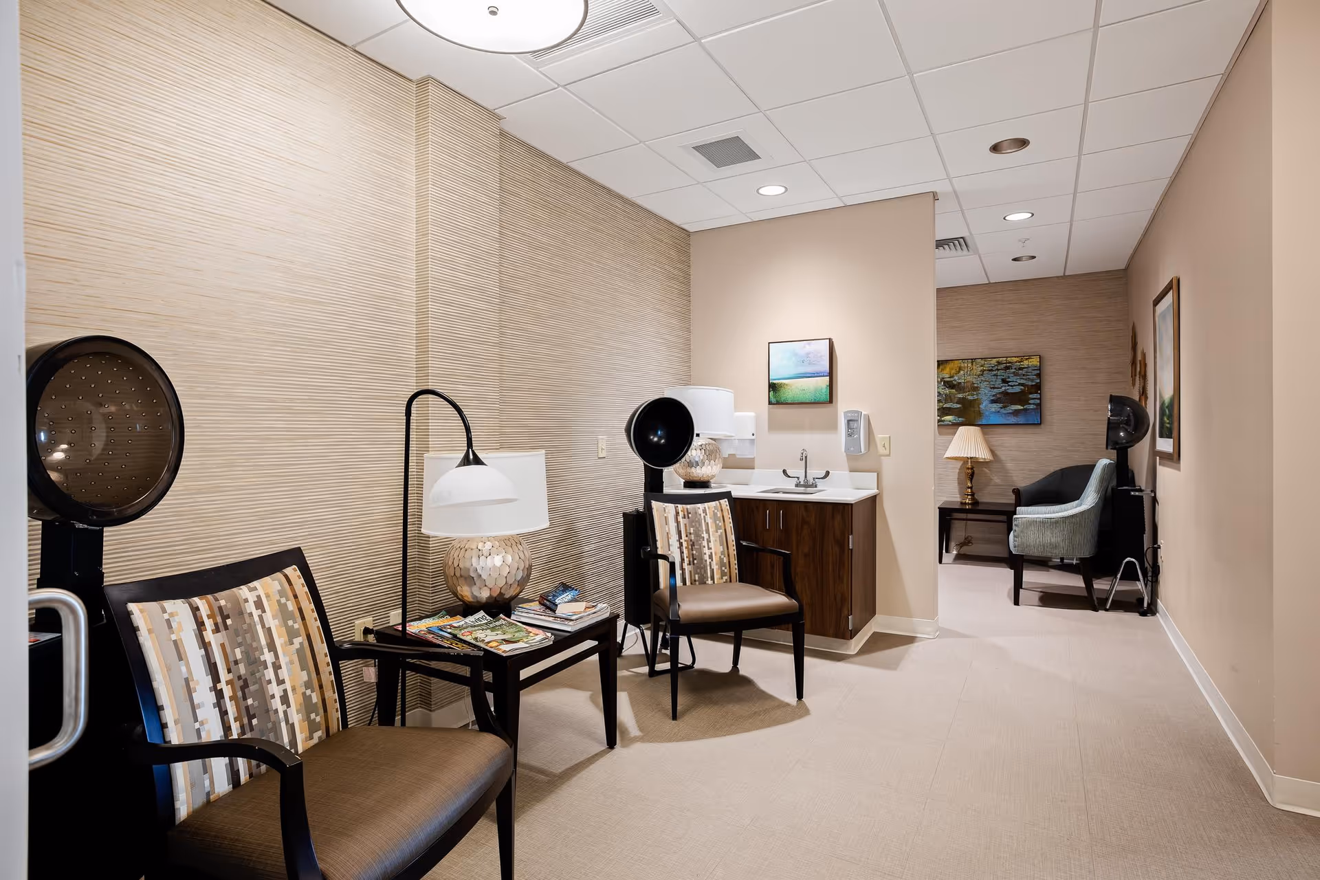 Small salon-style interior with chairs, hooded hair dryers, a sink countertop, side tables with lamps and magazines, and artwork on the walls.