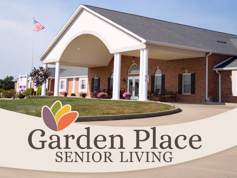 Exterior view of Garden Place Senior Living facility showing a brick building with white columns at the entrance, a curved driveway, green lawn, potted flowers, and an American flag on a flagpole.