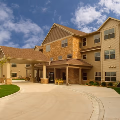 Exterior view of a multi-story senior living facility building with a covered entrance, beige siding, and brick accents under a partly cloudy blue sky.
