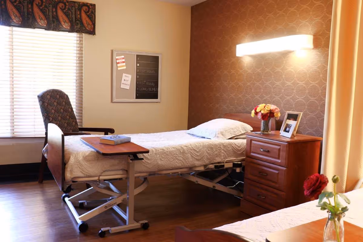 A well-lit bedroom in a senior living facility featuring a hospital-style bed with white bedding, a wooden nightstand with a vase of flowers and a framed photo, a comfortable upholstered chair near a window with blinds and a patterned valance, and a wall-mounted light fixture above the bed.