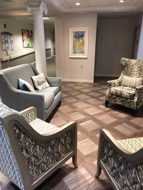 A cozy seating area in a senior living facility with a gray loveseat, two patterned armchairs, and a floral upholstered armchair arranged on a wood-patterned floor. The walls are light-colored with framed artwork and a white column visible in the background.