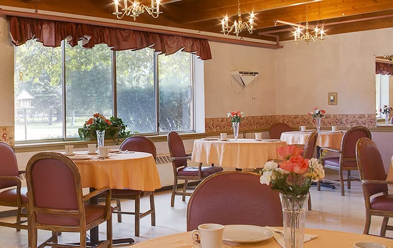 A dining room with round tables covered in beige tablecloths, each set with white cups and plates. The chairs have maroon upholstery and wooden frames. There are flower vases with pink and white flowers on the tables. Large windows with a view of green trees and a birdhouse outside let in natural light. The ceiling has wooden beams and chandeliers with multiple lights.
