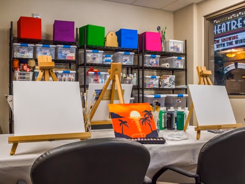 A creative art room setup with three wooden easels holding blank canvases and one canvas with a painted sunset scene featuring palm trees. Behind the easels are shelves filled with colorful storage boxes and clear plastic containers holding various art supplies. Two black chairs are positioned in front of the table with the easels. A window on the right side shows a sign that reads 'THEATRE' and 'NOW SHOWING'.