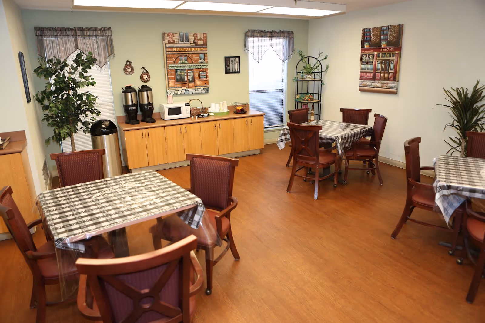 A dining room with several tables covered with checkered tablecloths and surrounded by wooden chairs with cushioned seats. There are two windows with valance curtains, a wooden cabinet holding coffee dispensers, a microwave, cups, and a basket. The room has light-colored walls decorated with framed pictures and plants in the corners.