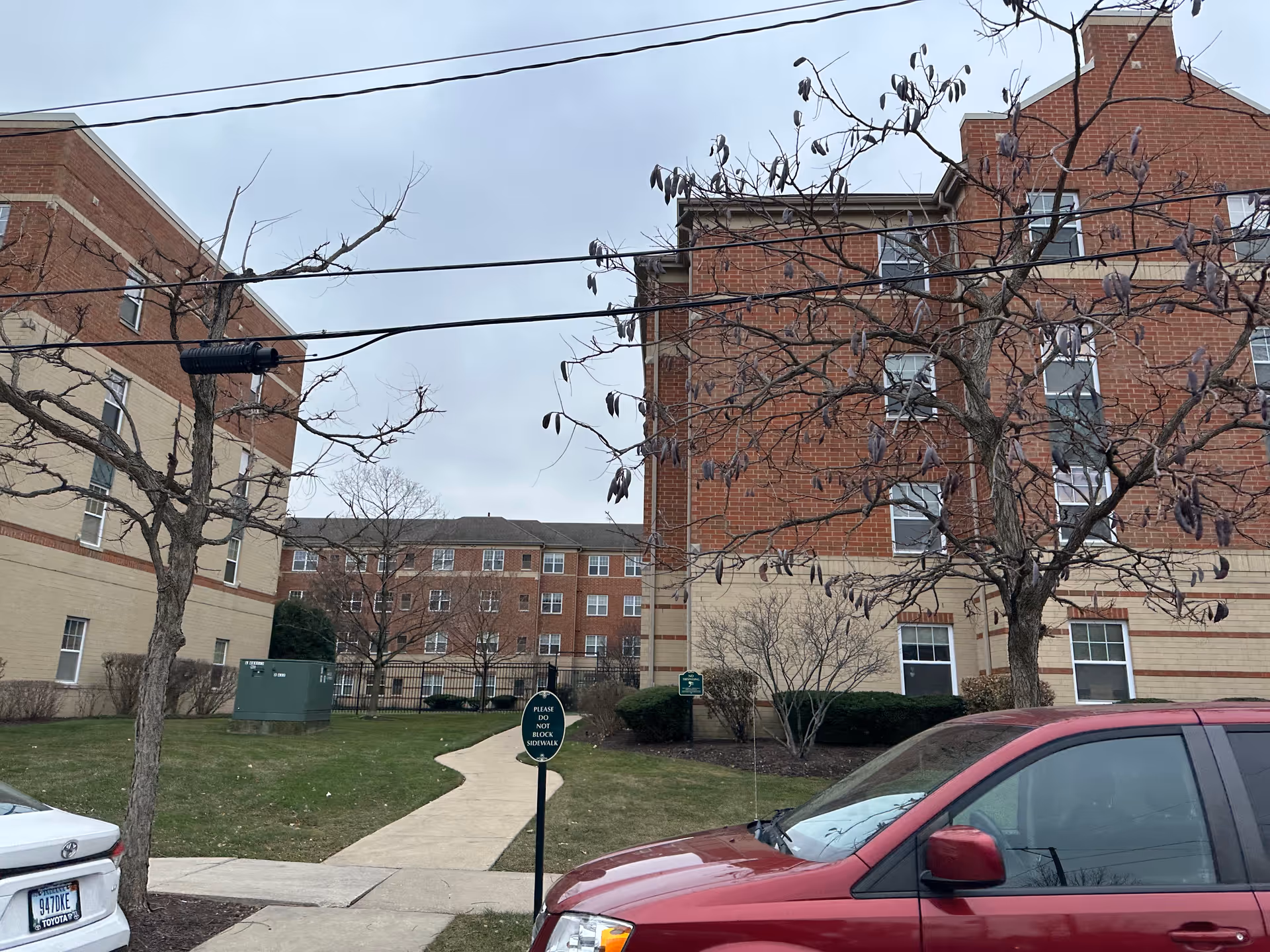 View of an outdoor area between brick buildings at Smith Village, showing a sidewalk, leafless trees, parked cars, and a sign that reads 'Please Do Not Block Sidewalk'.