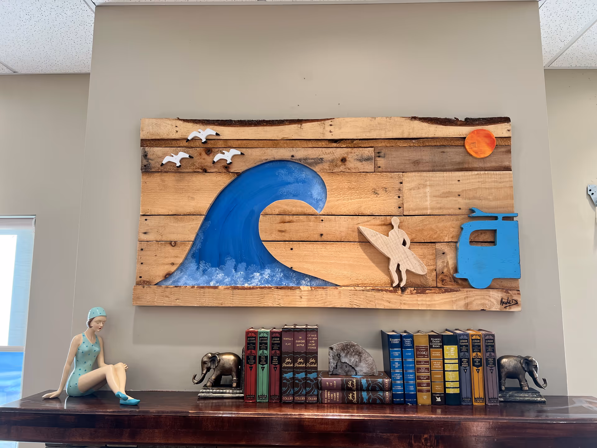 A decorative wooden wall art featuring a large blue wave, three white birds, an orange sun, a surfer holding a surfboard, and a blue van. Below the artwork is a dark wooden shelf with a figurine of a woman in a blue swimsuit and swim cap sitting, two elephant bookends, and several books arranged between the bookends.