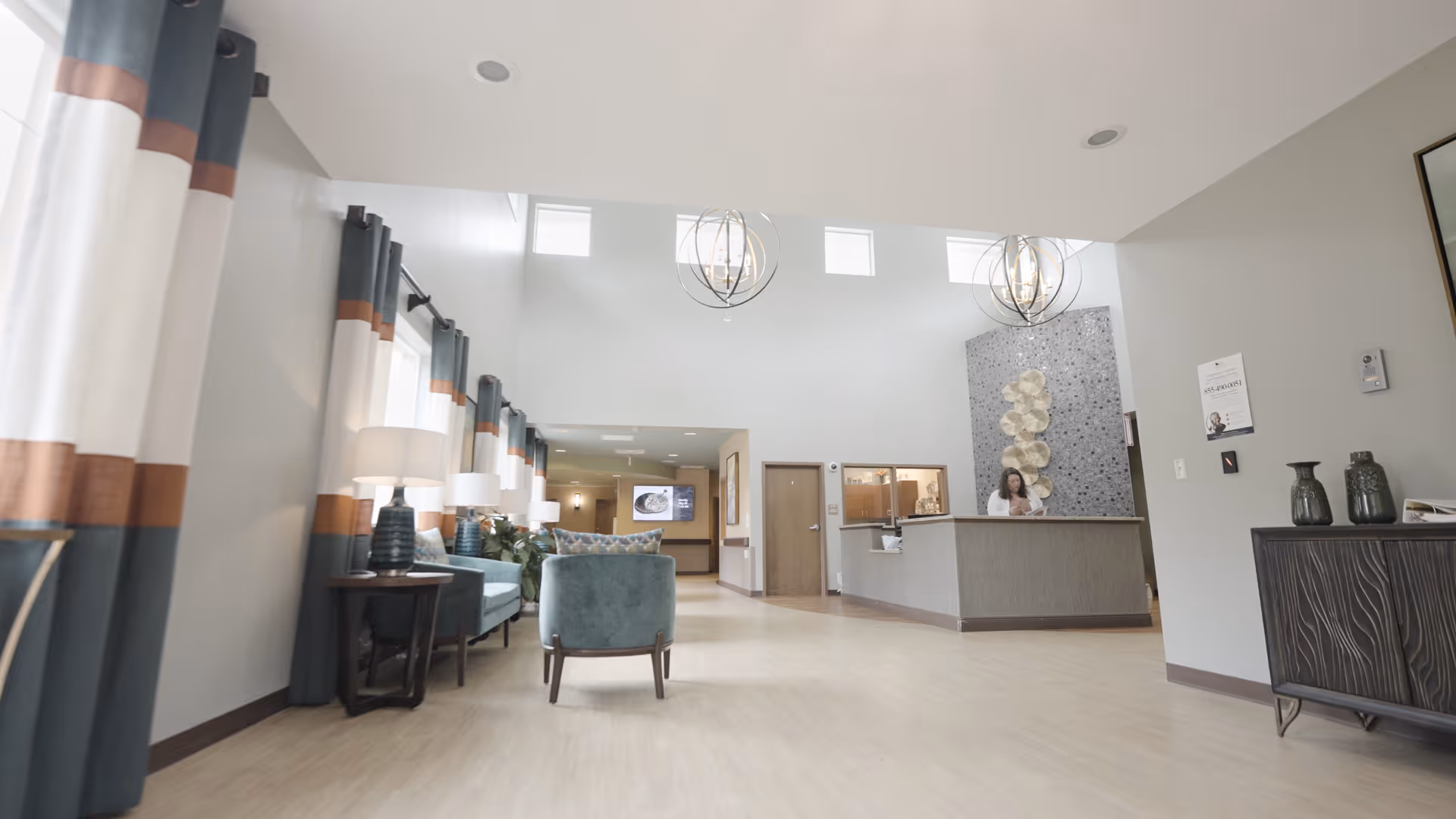 A spacious and well-lit reception area in a senior living facility with high ceilings and large windows near the ceiling. The room features teal and brown curtains, teal upholstered chairs, side tables with lamps, and a reception desk with a staff member behind it. Decorative wall art and modern light fixtures hang from the ceiling.