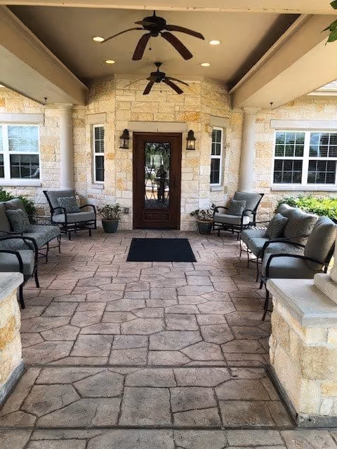 Covered outdoor seating area with cushioned chairs arranged along stone walls, ceiling fans overhead, and a wooden door with glass panels at the far end, flanked by two windows and wall-mounted lantern lights.