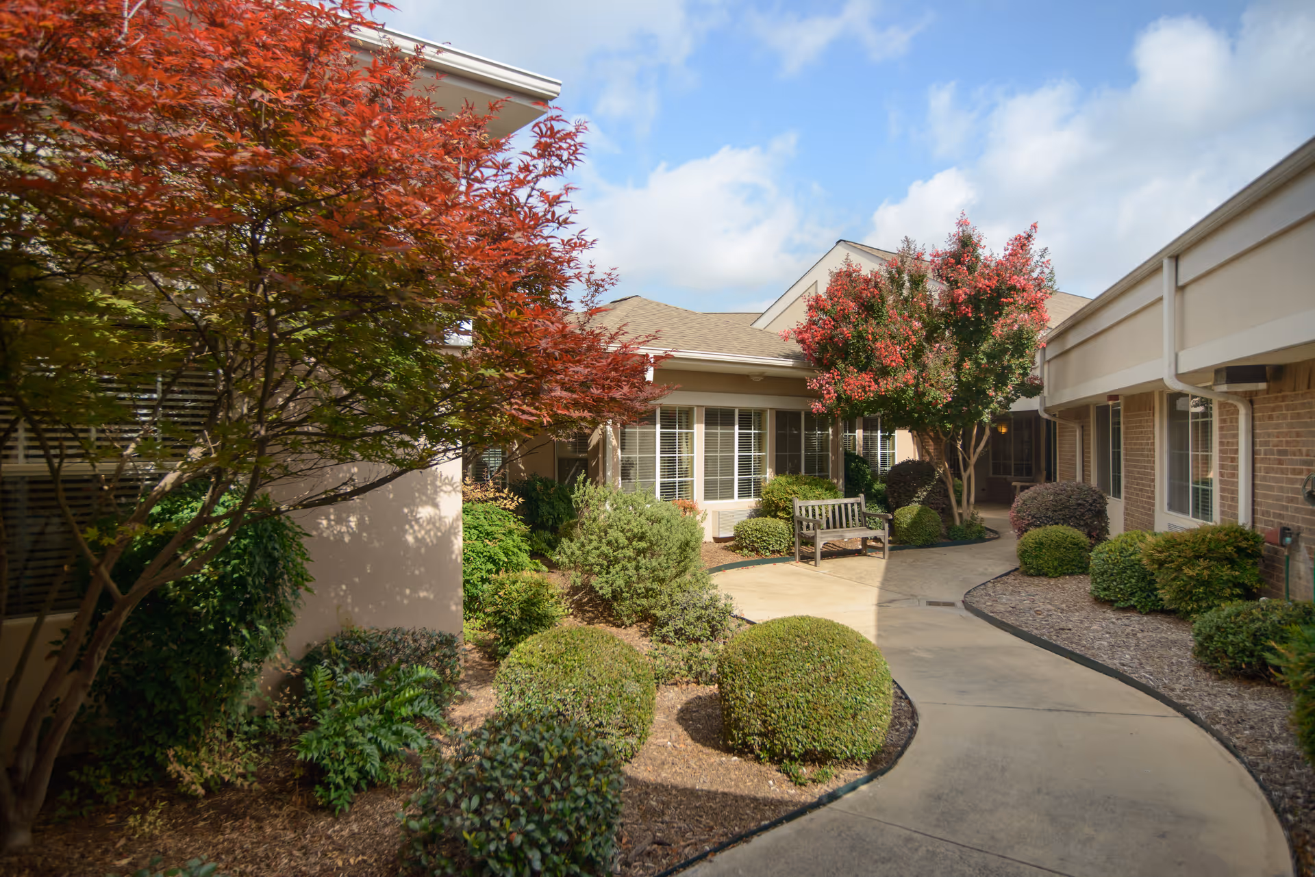 A landscaped outdoor courtyard area at a senior living facility with a curved concrete walkway, neatly trimmed bushes, a wooden bench, and trees with red and green leaves. The building exterior features beige walls and windows with white blinds under a partly cloudy sky.